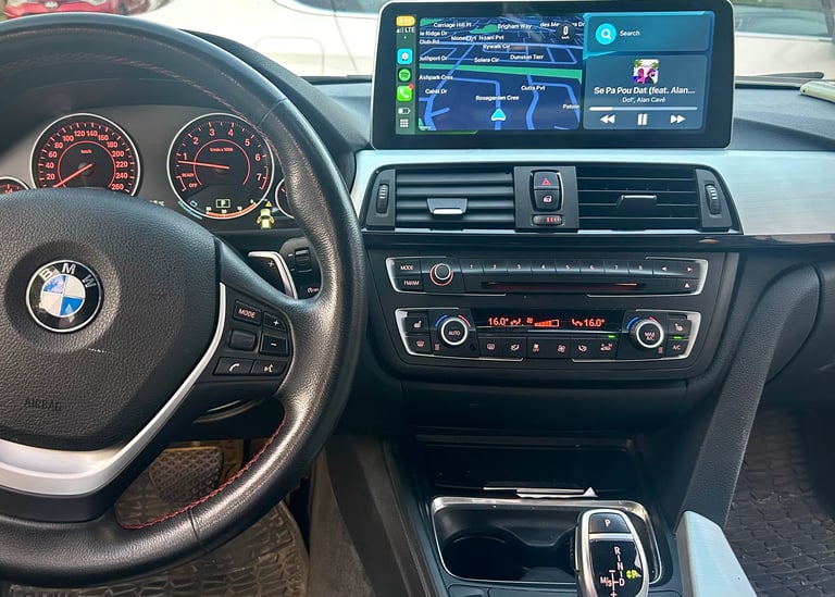 Interior of a BMW car showing steering wheel with logo, analog gauges, touchscreen infotainment system displaying navigation, and climate controls