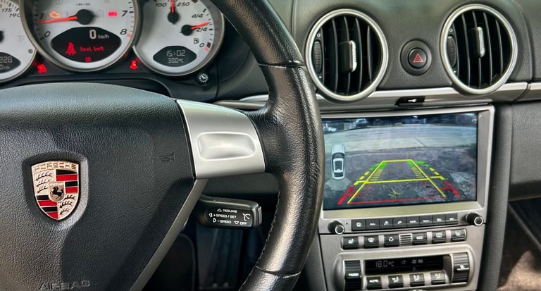 Porsche car interior showing steering wheel with logo, dashboard gauges, and touchscreen display with rear-view camera feed